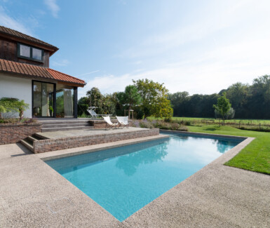 Azuurblauw moaïek zwembad in L-vorm aangelegd door RVS SWIMMINGPOOLS in tuin in Destelbergen